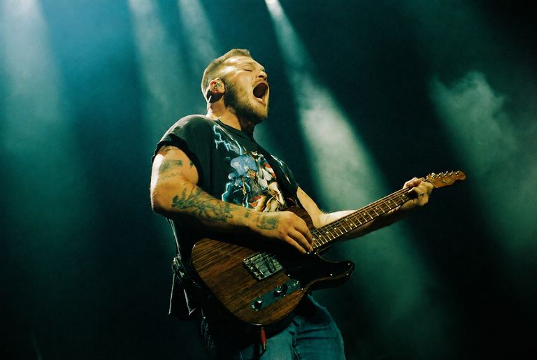 Country music artist Zach Bryan singing passionately while playing electric guitar live under dramatic stage spotlights.